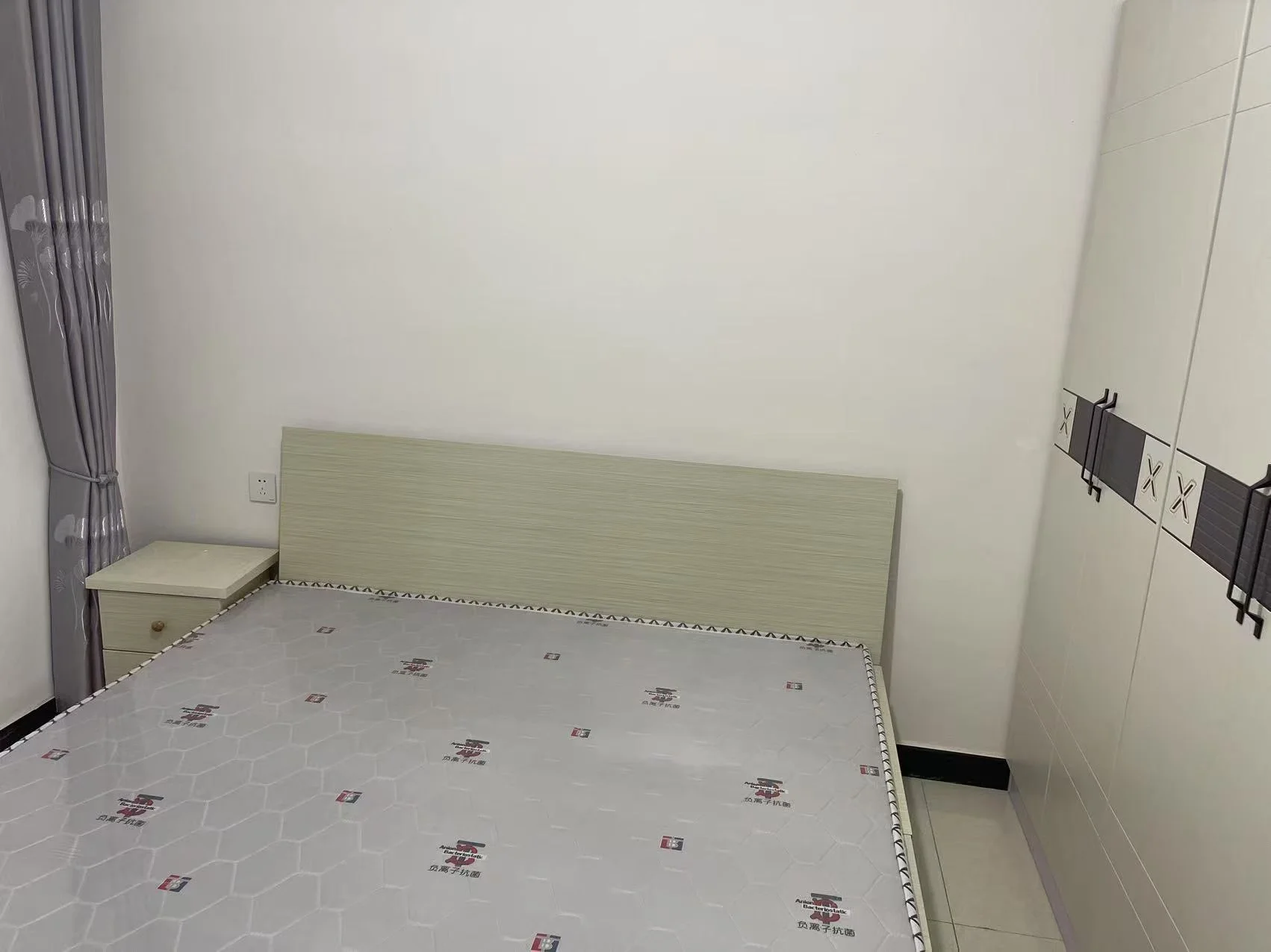 卧室空间，床铺为深色木质床架，搭配带有几何图案的床品。房间墙面简洁，采光适中，整体呈现基础且整洁的居住环境。