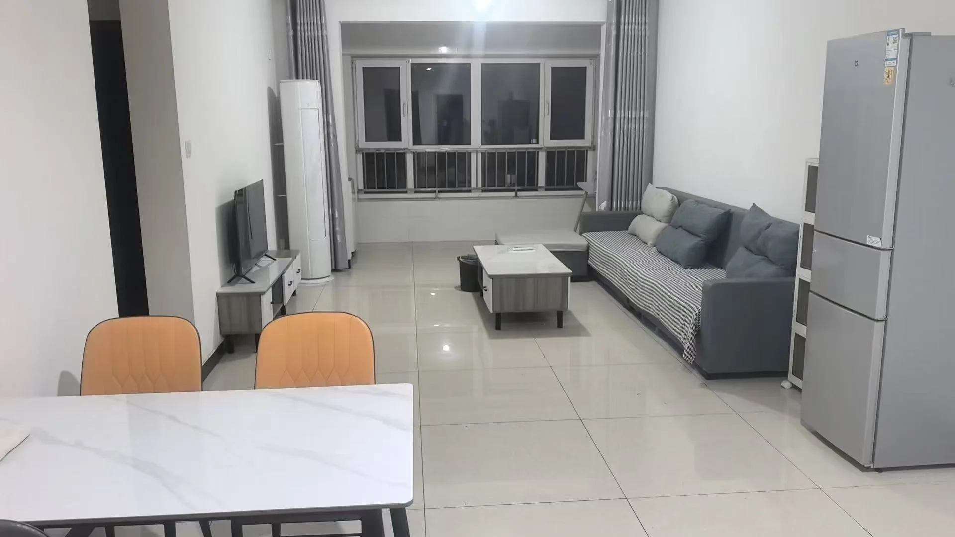 这是一个开放式起居空间，地面铺设浅色瓷砖，配有简约风格的沙发和餐桌区域。房间采光良好，整体装修简洁明亮，具备基础的居住功能。