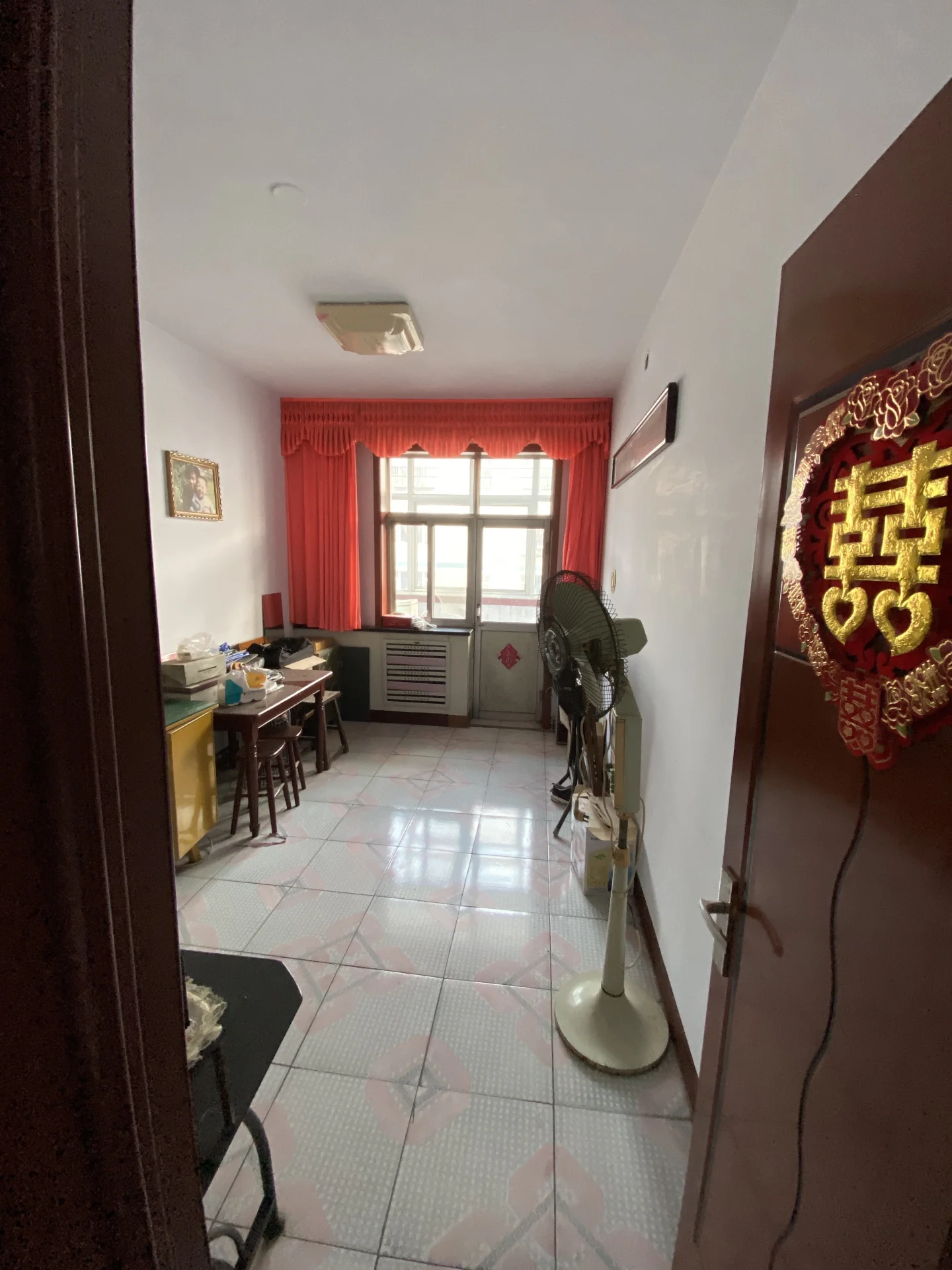 这是一个功能性较强的室内空间，地面铺设浅色瓷砖，配有窗户和空调设备。房间内摆放着办公桌椅和一台大型风扇，整体装修风格偏向实用简约，光线适中。