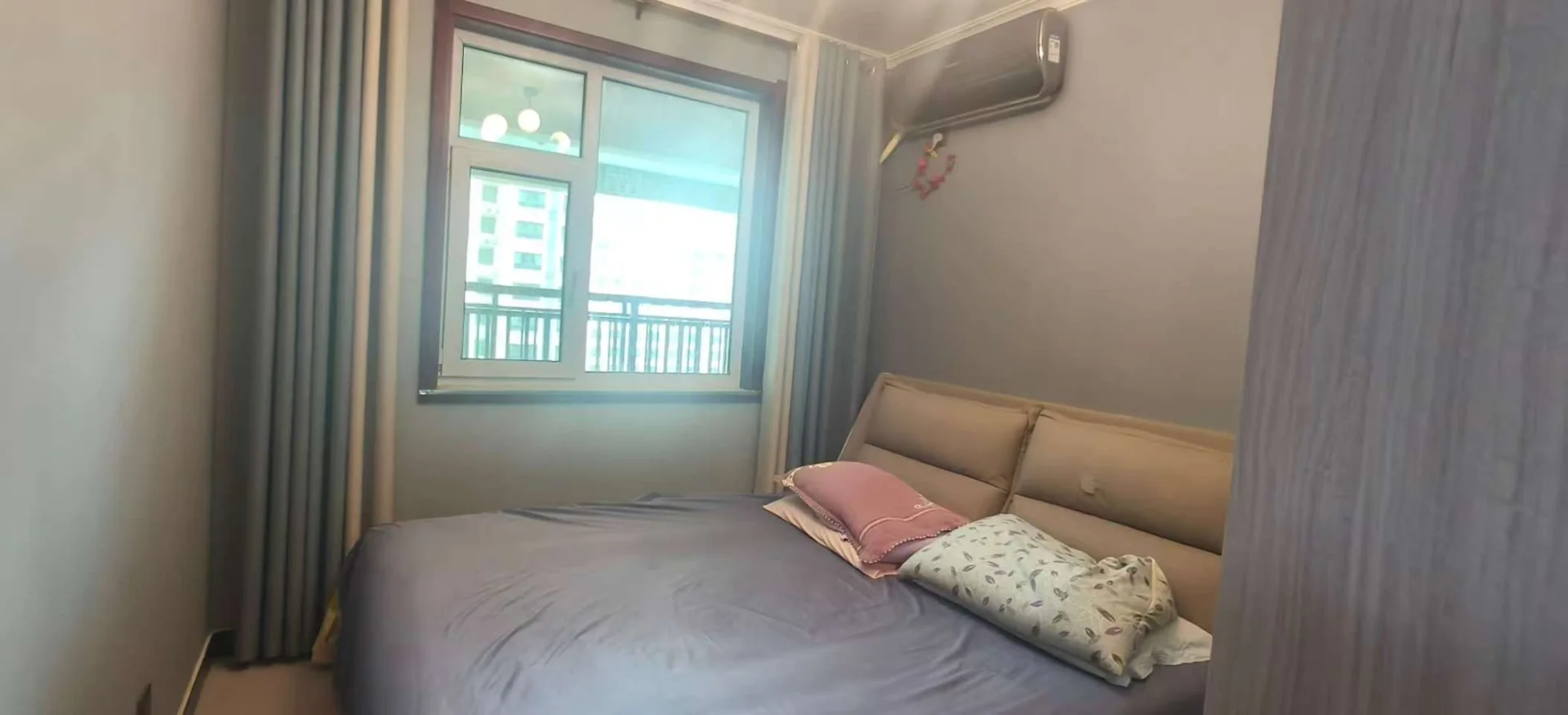 卧室空间，床铺为米色软包设计，搭配简约的床品和枕头。房间采光良好，窗外可见城市建筑轮廓，整体呈现简洁舒适的现代风格。