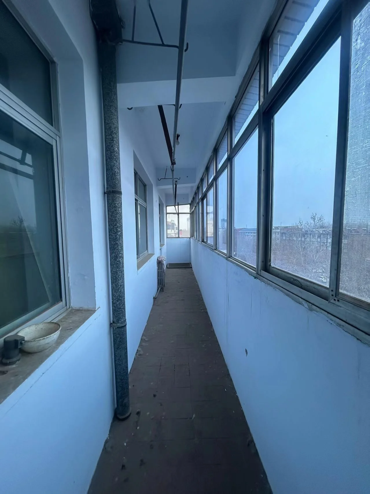 建筑走廊场景，墙面为白色，地面铺设砖块，配有大型窗户和裸露的管道结构。整体环境较为简朴实用，光线适中。