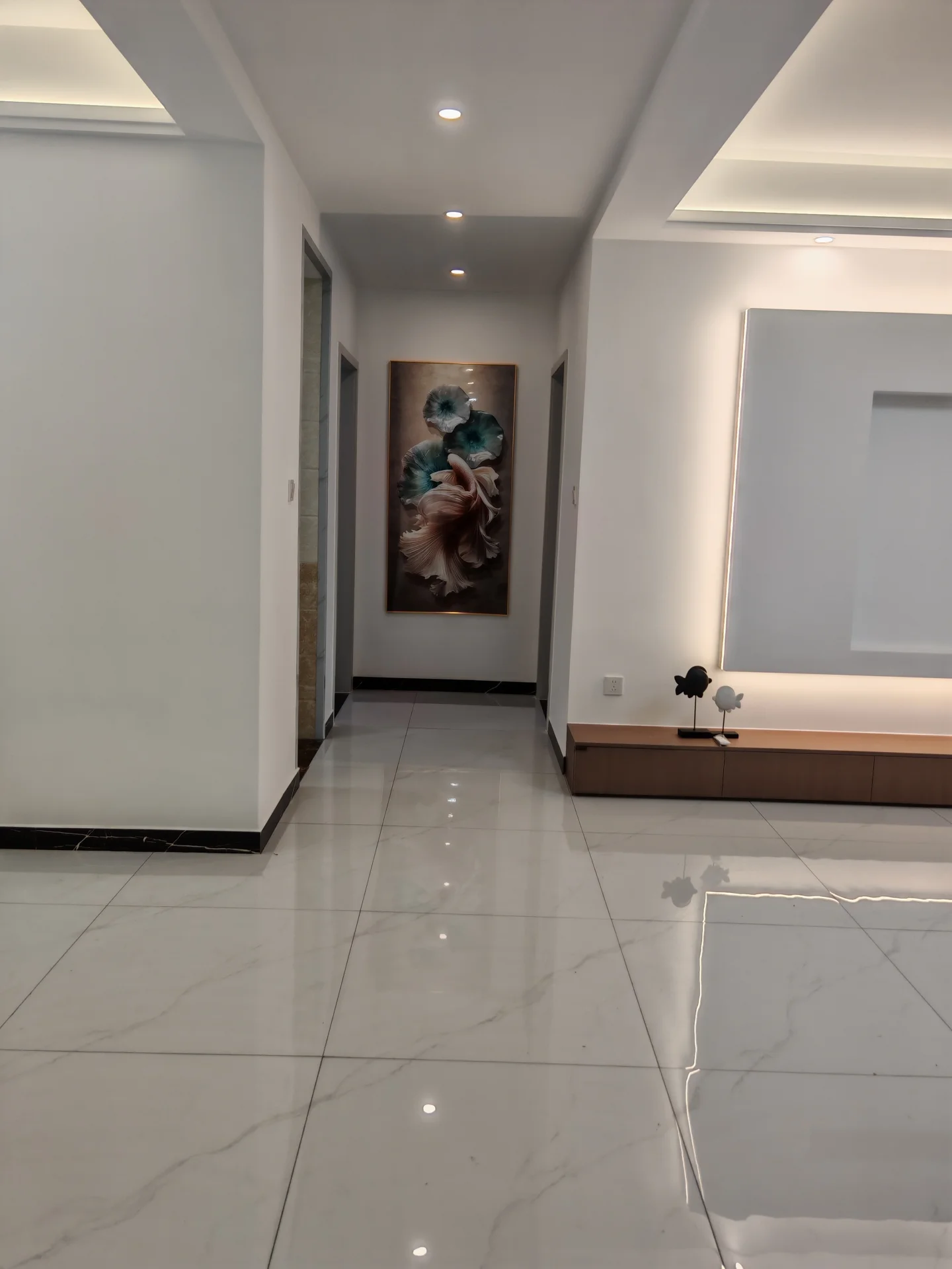 走廊空间，地面铺设大尺寸白色大理石瓷砖，墙面简洁，配有现代简约的灯光设计和一幅大型艺术画作。整体环境明亮、整洁，具有现代商业或公共区域的风格。