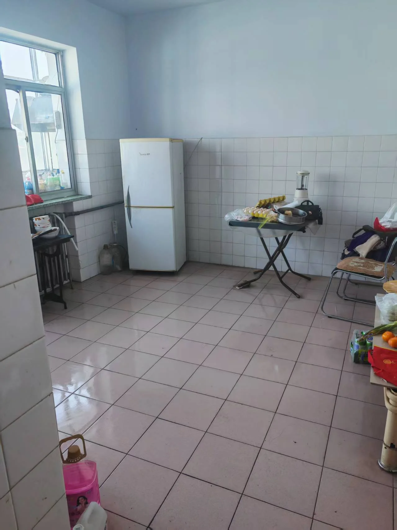 厨房空间，地面铺设浅色瓷砖，墙面为白色瓷砖，配有大型白色冰箱和操作台。整体环境简洁明亮，属于基础功能型装修。