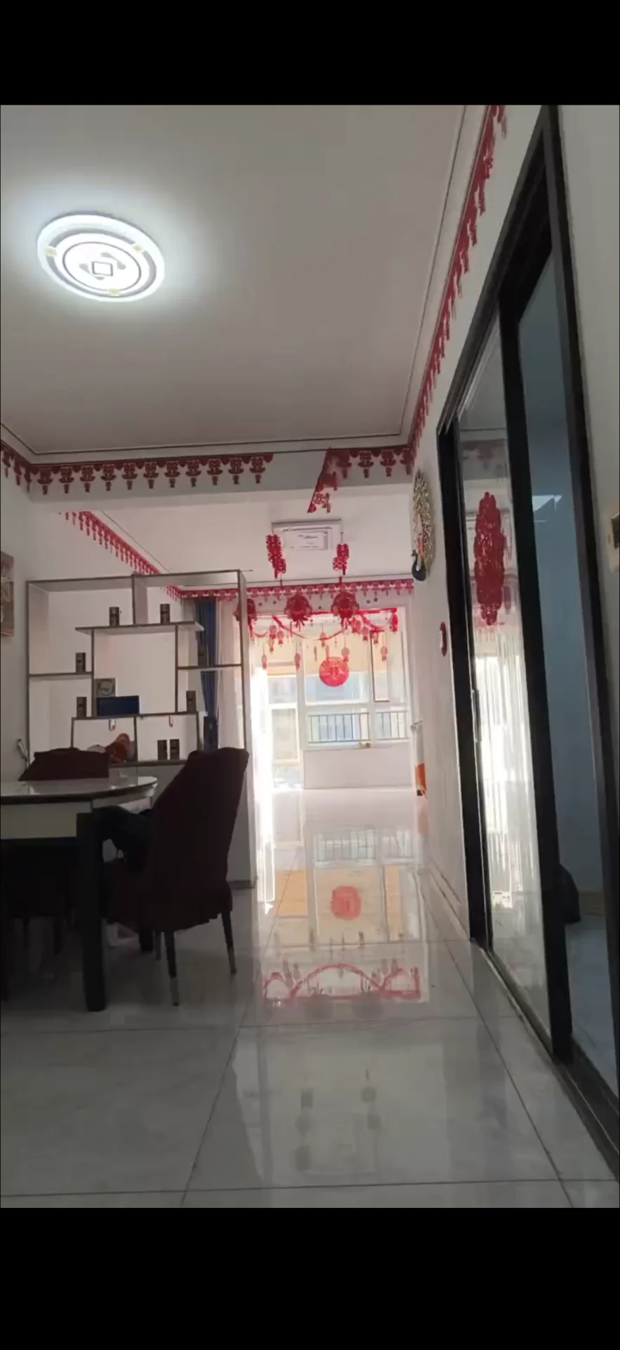 这是一个宽敞的室内空间，地面铺设浅色瓷砖，天花板配有圆形吸顶灯。房间内摆放着餐桌和深色沙发，整体装修风格偏向基础实用型，光线充足。