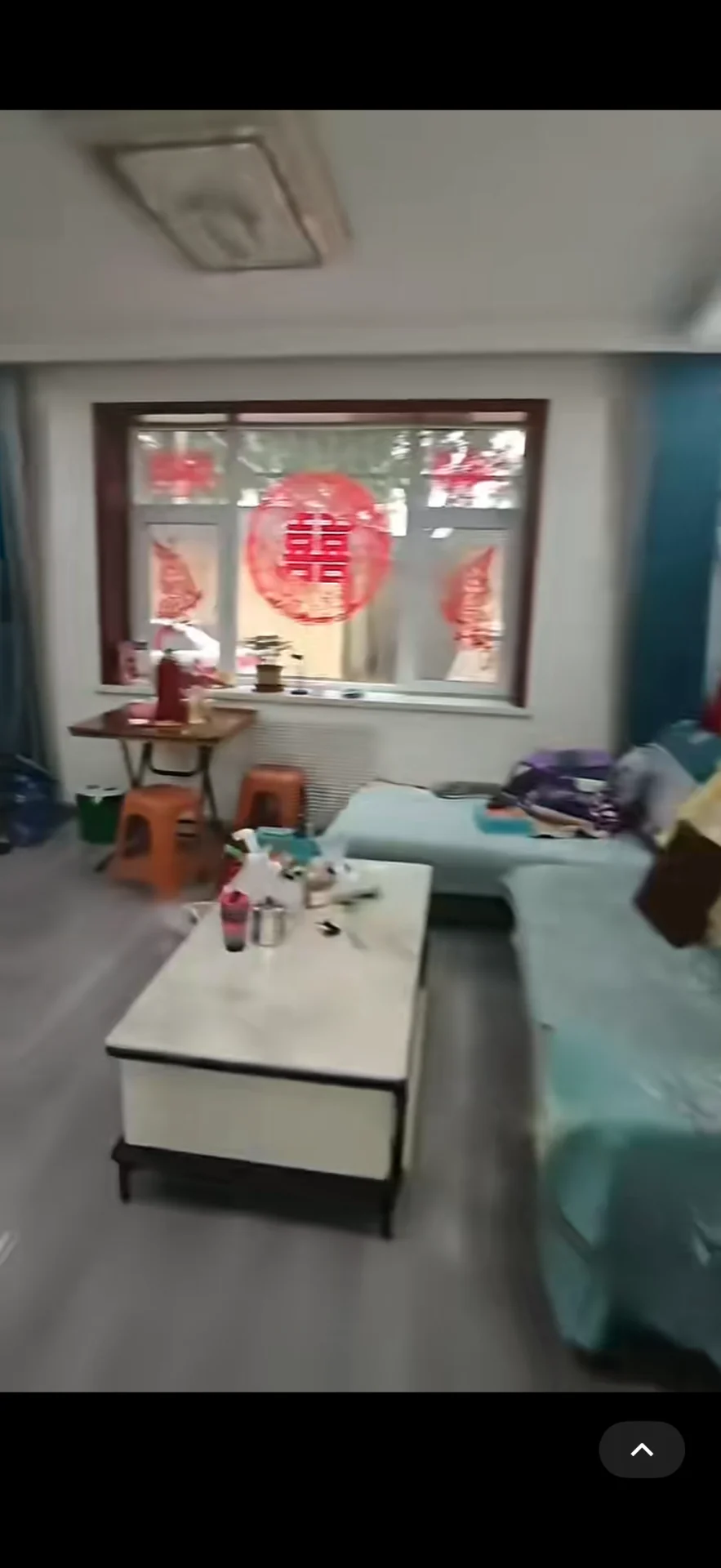 这是一个室内空间，地面为浅色瓷砖，房间内摆放着一张白色大方桌和几张床铺。整体装修风格较为基础，采光适中，呈现出一种功能性居住环境的氛围。