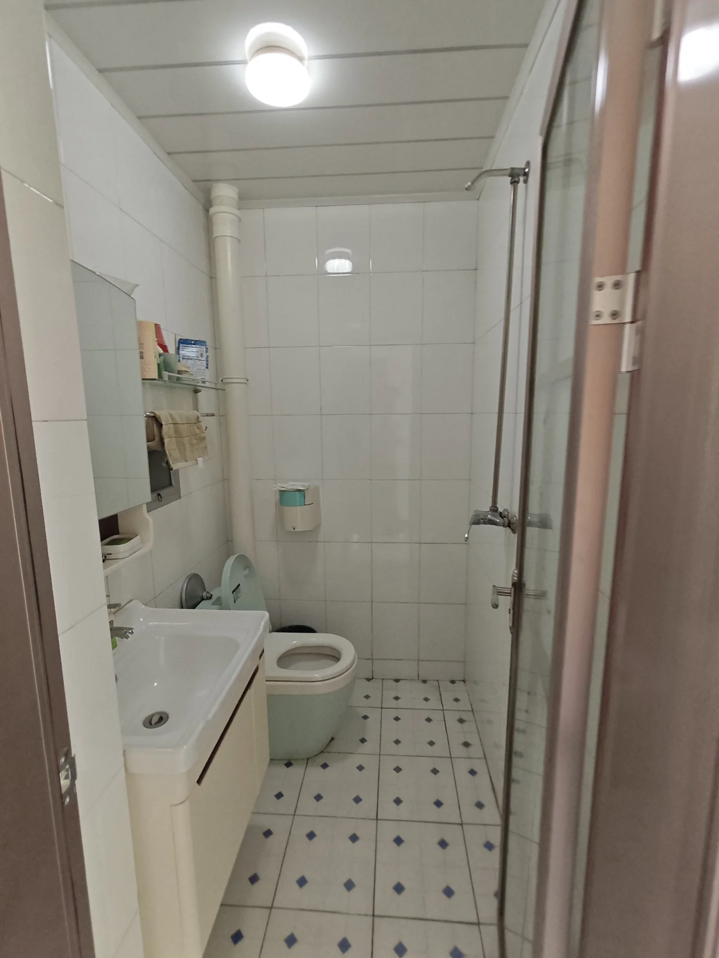 卫生间空间，墙面和地面铺设白色及浅蓝色瓷砖，配有白色洗手台、马桶和玻璃淋浴房。光线明亮，整体装修风格简洁实用。