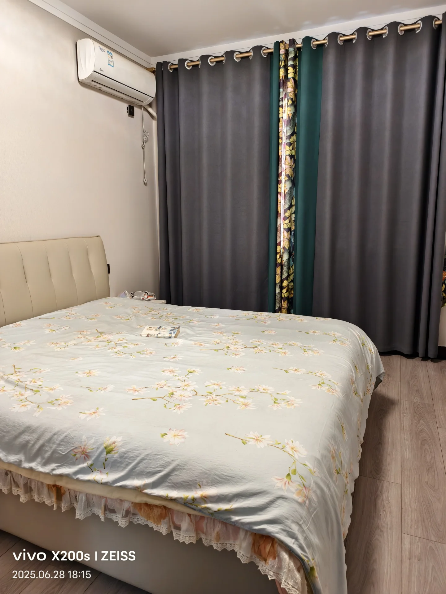 卧室空间，床铺采用白色系带有花卉图案的床单，搭配米色软包床头板。房间内安装有空调，整体装修风格偏向温馨舒适的现代简约风，采光适中。
