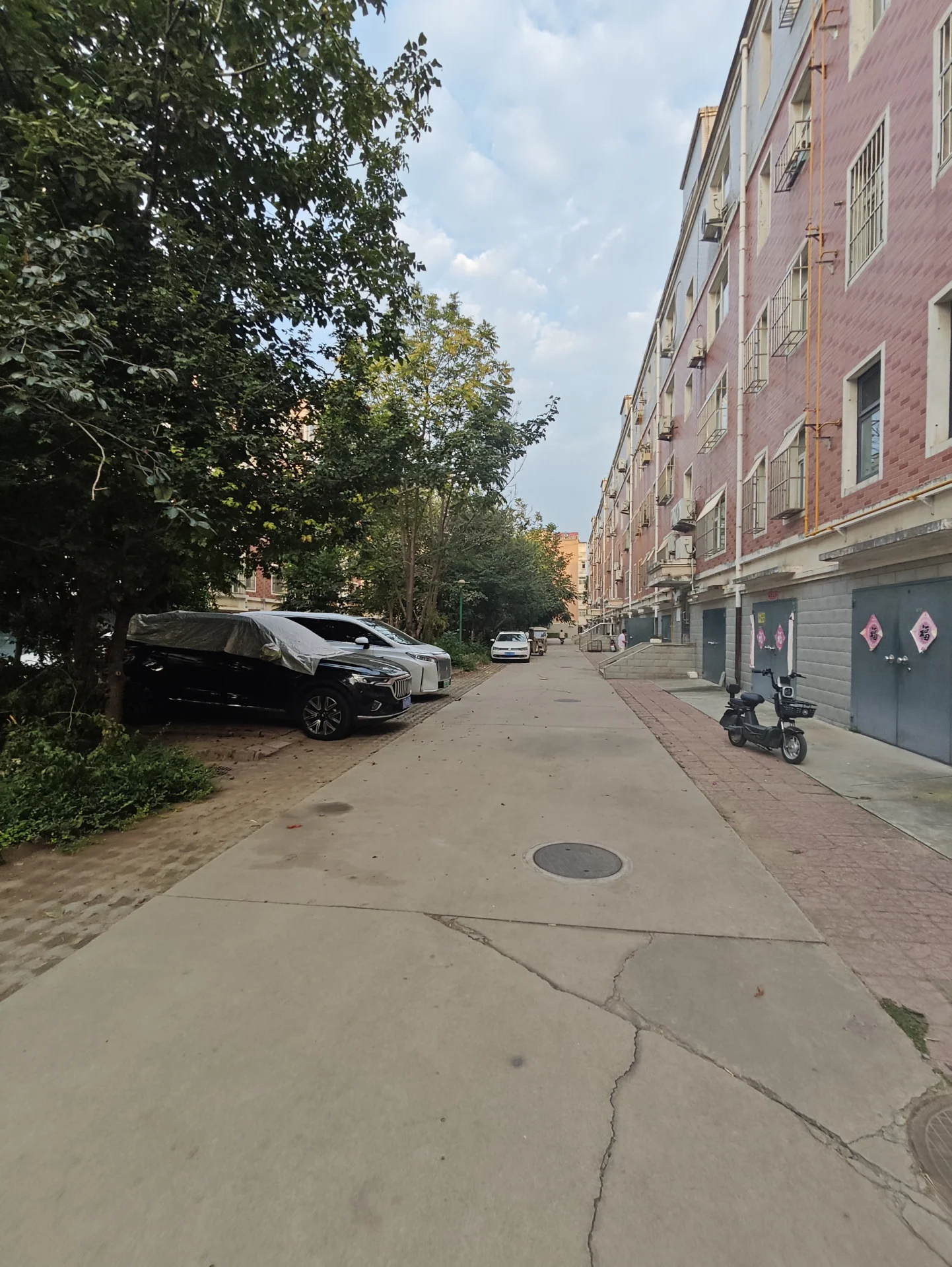 一条城市街道，两侧是多层住宅建筑，道路中央有路面井盖，绿化带分布在左侧，整体呈现典型的城市居住环境。