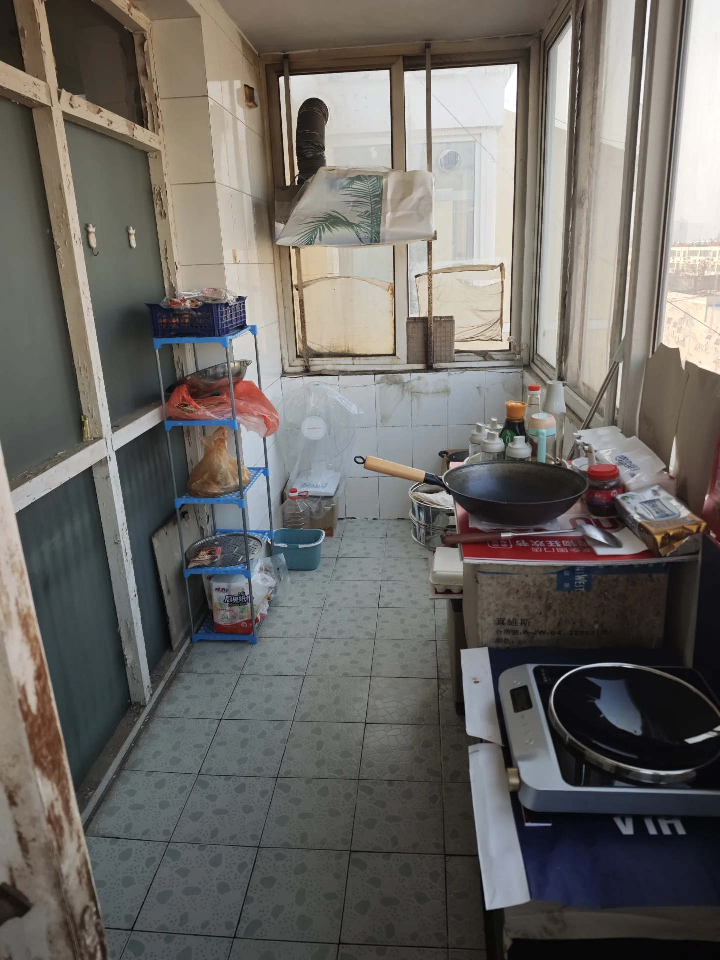 这是一个带有大窗户的室内空间，地面铺设浅色瓷砖，墙面为浅色调。空间内摆放着一个金属置物架，上面堆放着食材和包装物品，右侧放置了烹饪设备和厨具。整体环境偏向功能性操作区域。