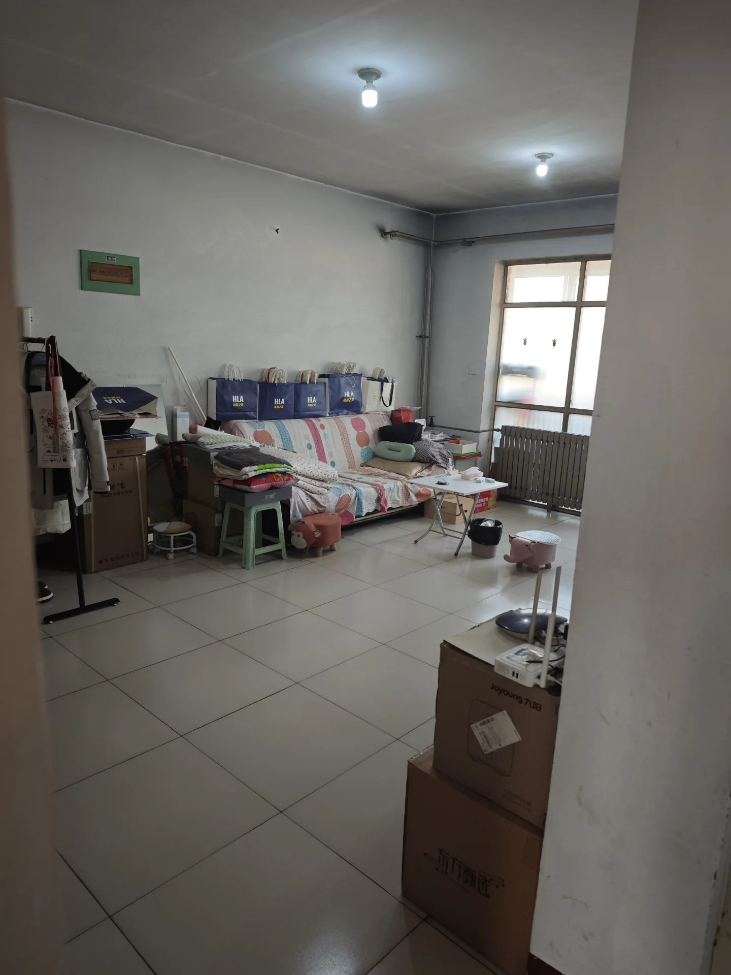 这是一个空旷的室内空间，地面铺设白色瓷砖，墙面为素色，整体装修风格基础简约。房间内摆放着床、衣架和一些箱子，采光适中，呈现出待布置或刚入住的居住环境。