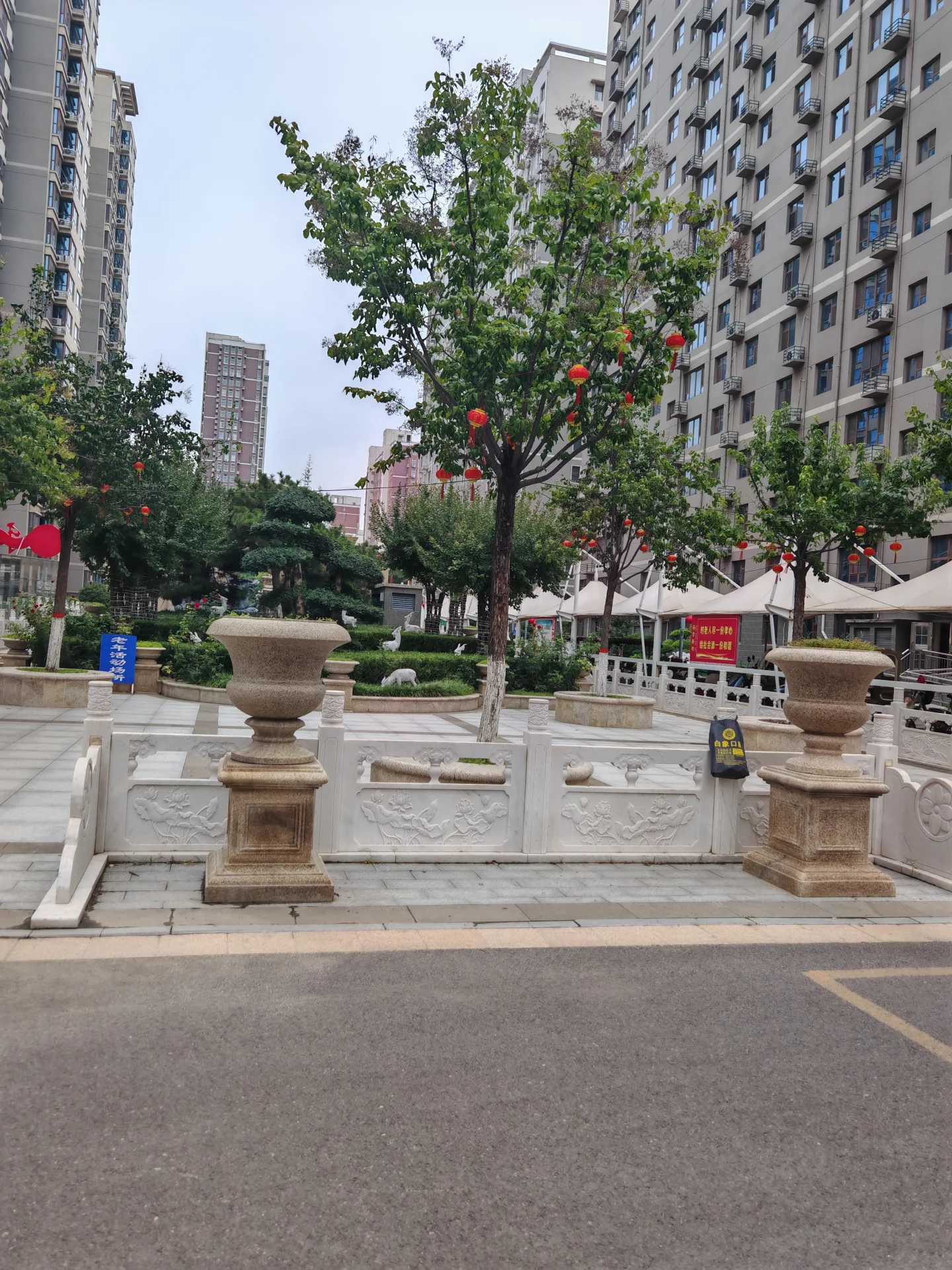 一棵枝繁叶茂的树位于街道中央，周围是现代高层住宅建筑的背景，树下设置有装饰性的石制围栏和红色灯笼，整体环境整洁有序。