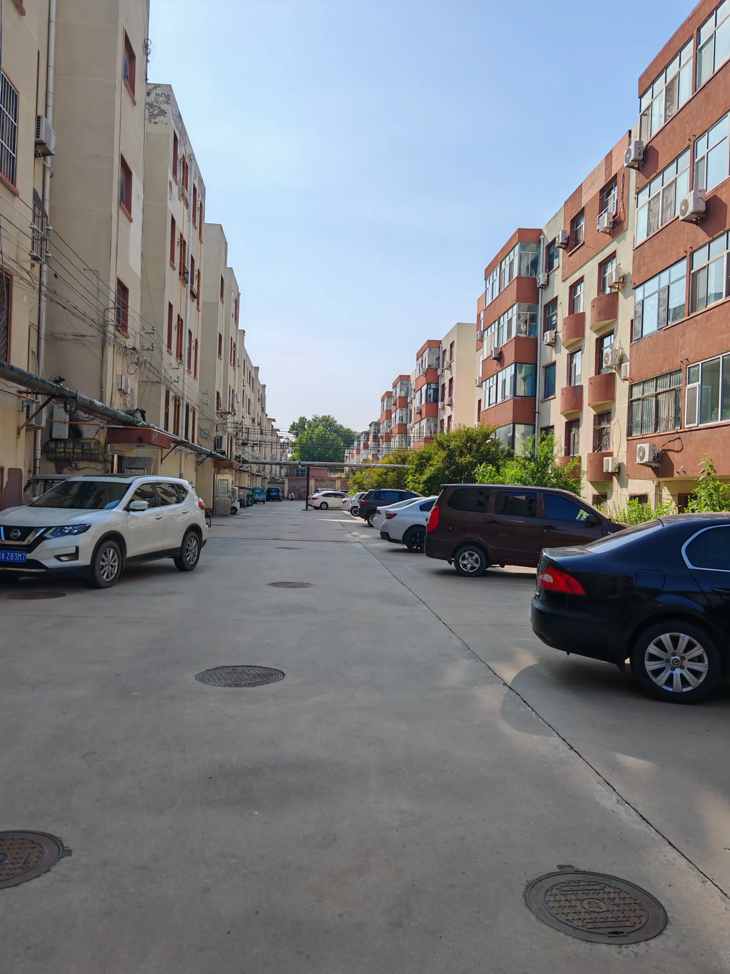 一条城市住宅区内的街道，两侧是多层砖红色住宅楼，建筑风格较为朴素和实用。街道宽敞，地面铺设水泥材质，停放着几辆私家车。整体环境展现了典型的城市居住场景。
