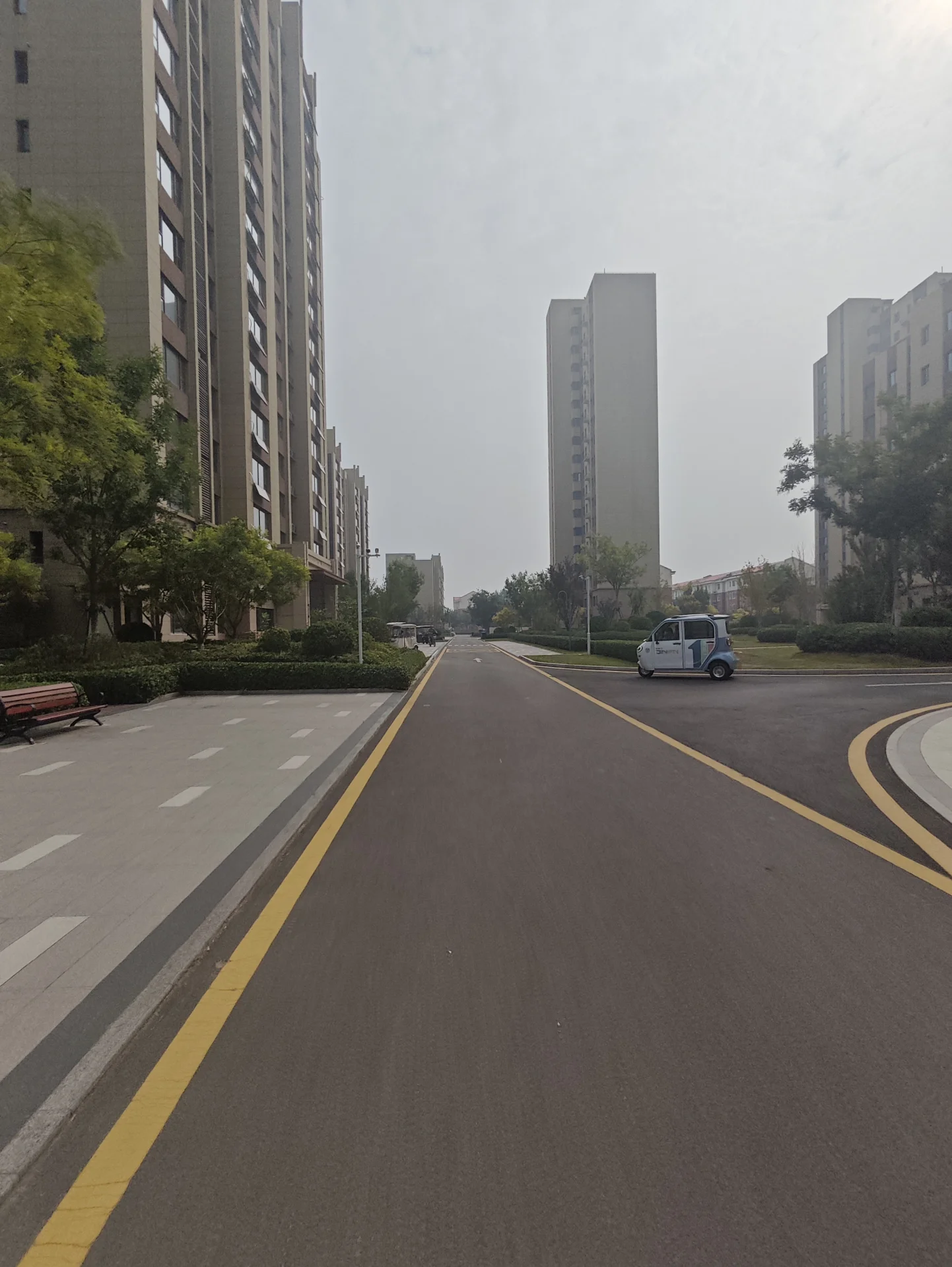一条城市住宅区内的道路，两侧是高层住宅建筑，道路平整，有清晰的交通标线和绿化带环绕，整体环境宁静有序。