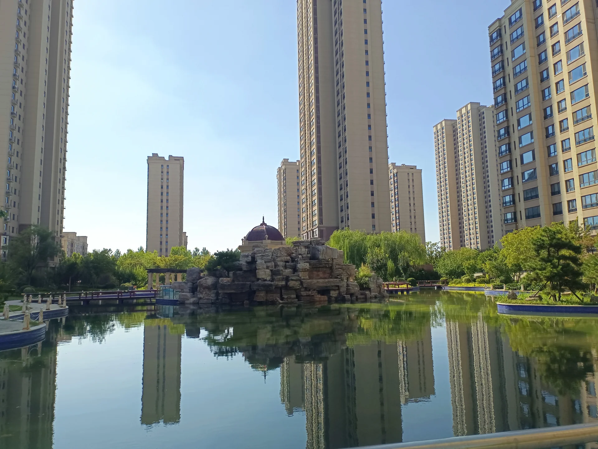 中心湖泊映照着周围高层住宅楼的倒影，湖边设有景观小岛和栈道，绿化覆盖良好，整体呈现现代城市公园的宁静氛围。