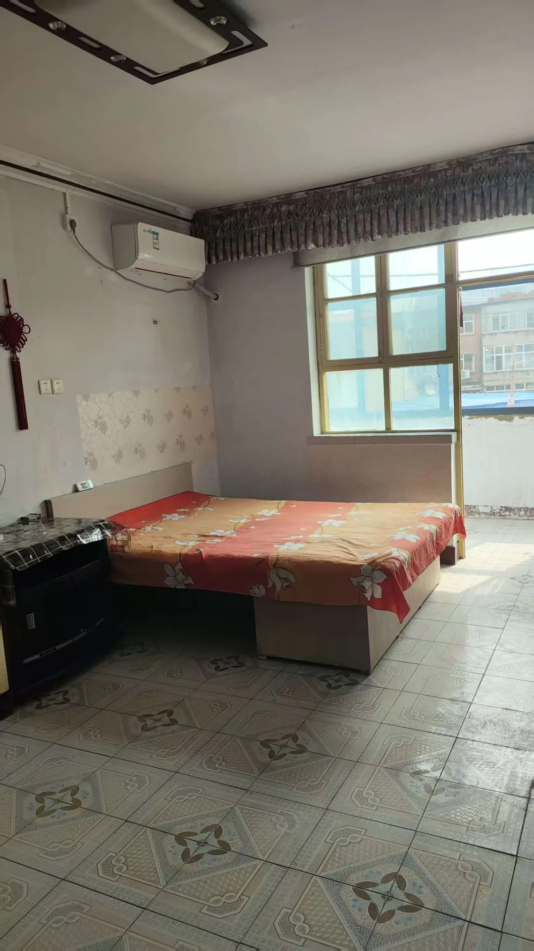 卧室空间，床铺配有花纹床单，房间内安装有空调和窗户。光线适中，整体呈现基础的居住环境。