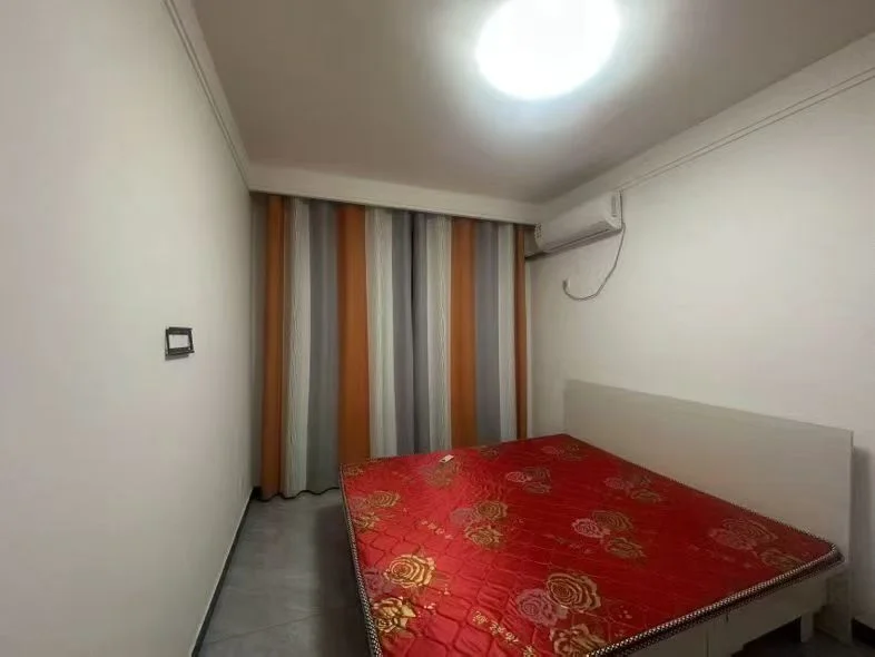 卧室空间，床铺为深红色大床，搭配简洁的白色床架。房间墙面为浅色调，采光适中，整体呈现基础且功能明确的居住环境。