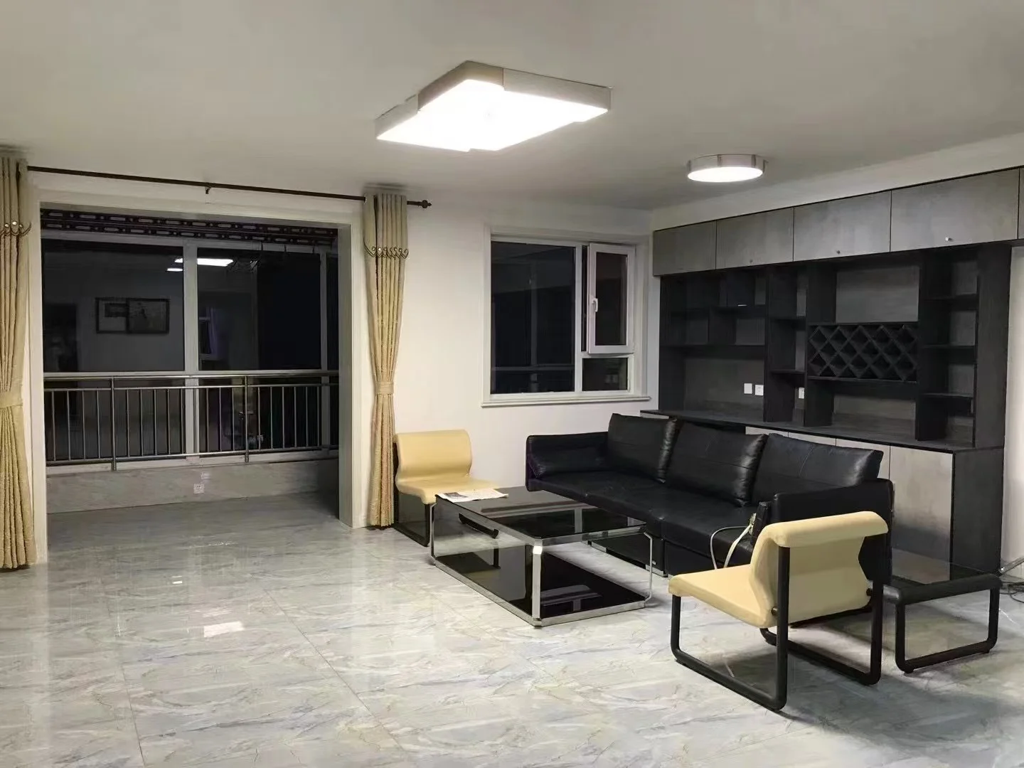 客厅空间，地面铺设大理石纹瓷砖，配有黑色皮质沙发和米色休闲椅。房间采光良好，整体装修风格偏向现代简约，空间开阔。