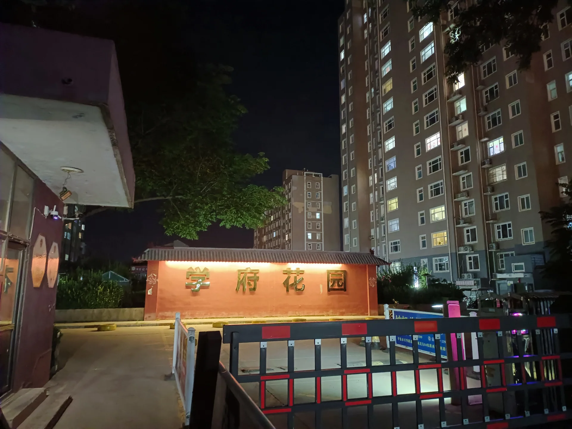 一座带有红色招牌的低层建筑位于高层住宅楼前方，夜间灯光亮起，整体呈现出社区入口或管理区域的氛围。