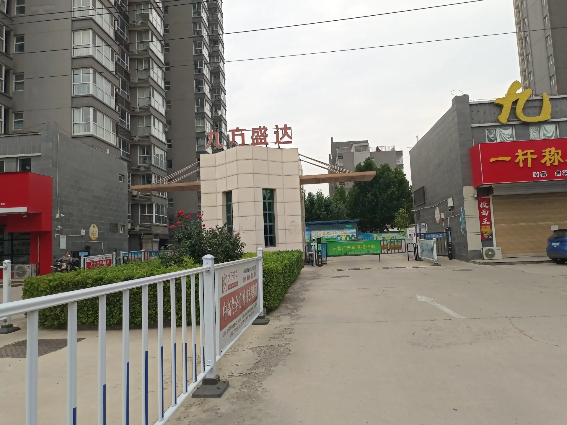一处住宅小区入口场景，主体建筑为多层现代风格的住宅楼，入口区域设有围栏和宣传标识。周边环境开阔，整体呈现城市居住区的氛围。
