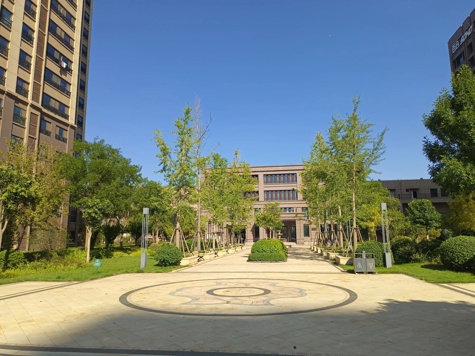 一栋多层现代风格的建筑位于中心，周围环绕着茂密的树木和开阔的广场区域。建筑立面简洁，整体环境绿化良好，呈现出宁静和谐的社区氛围。
