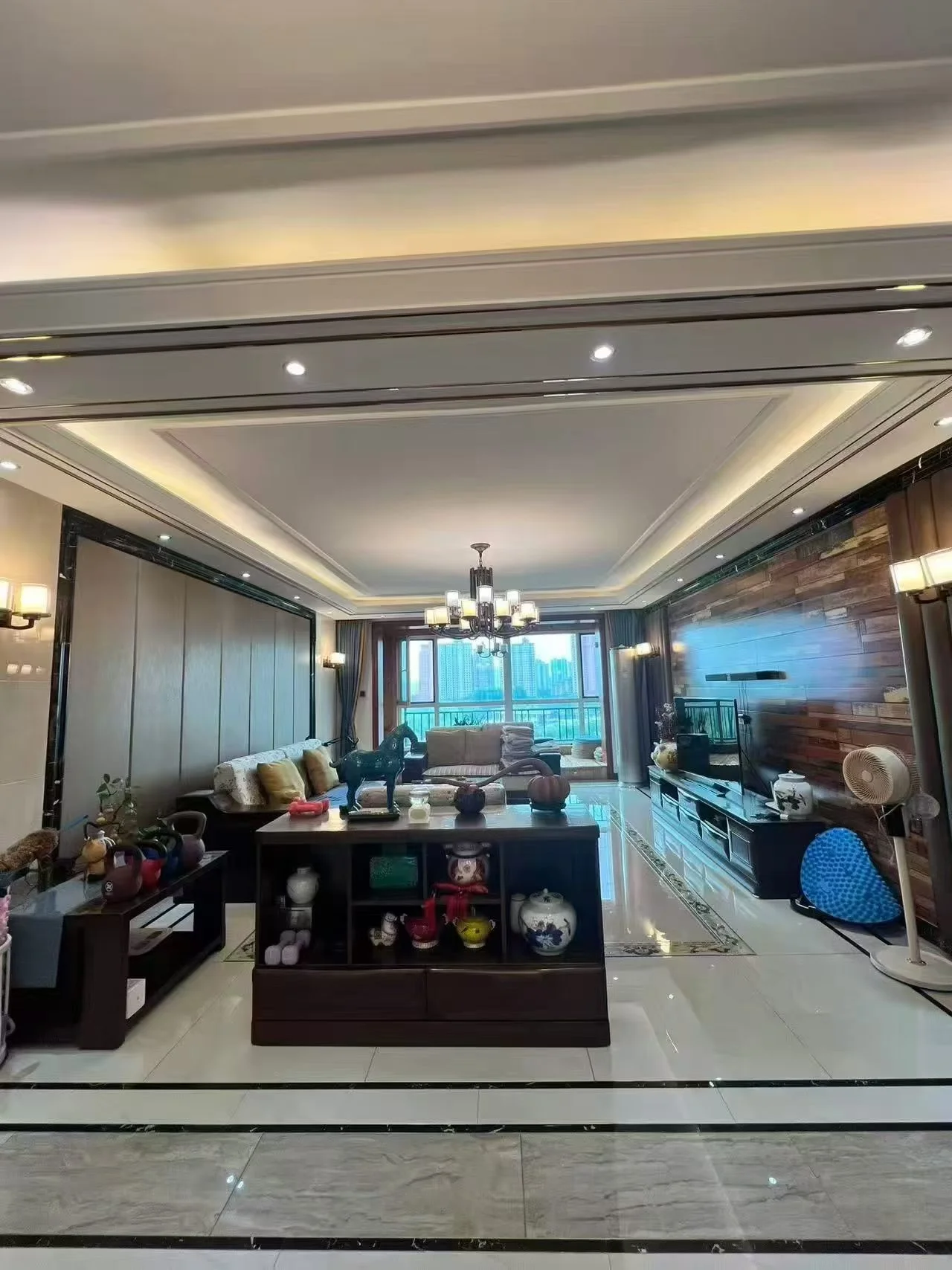这是一个宽敞的客厅空间，采用了深色木质家具和浅色大理石地面相结合的设计。天花板设计有层次感，配有吊灯照明，整体装修风格偏向传统或豪华型，采光良好。