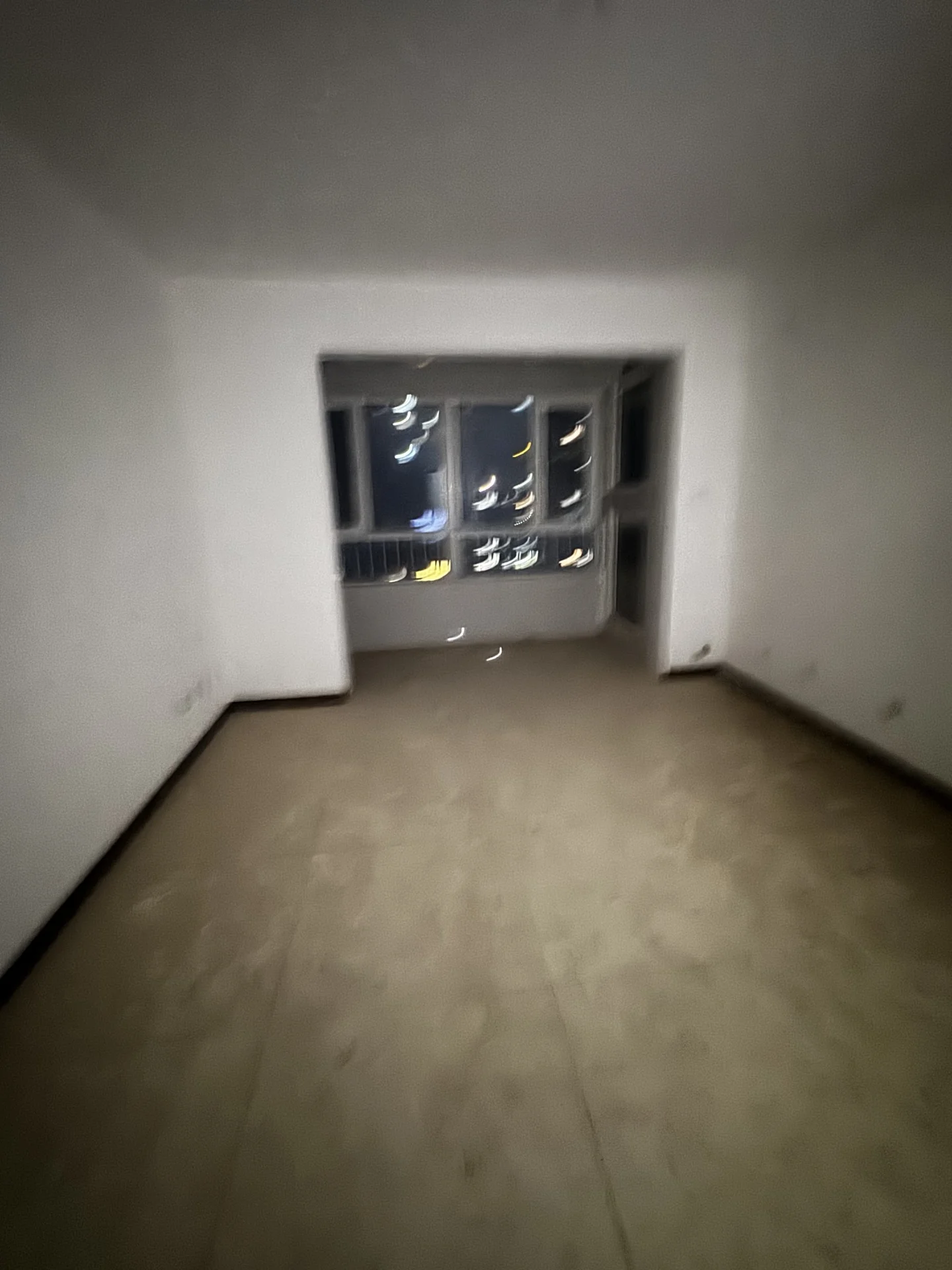 空旷的室内空间，墙面为白色，地面铺设浅色地砖或地板。房间结构简单，采光主要依赖远处的窗户，整体呈现出待装修或基础居住环境的特点。