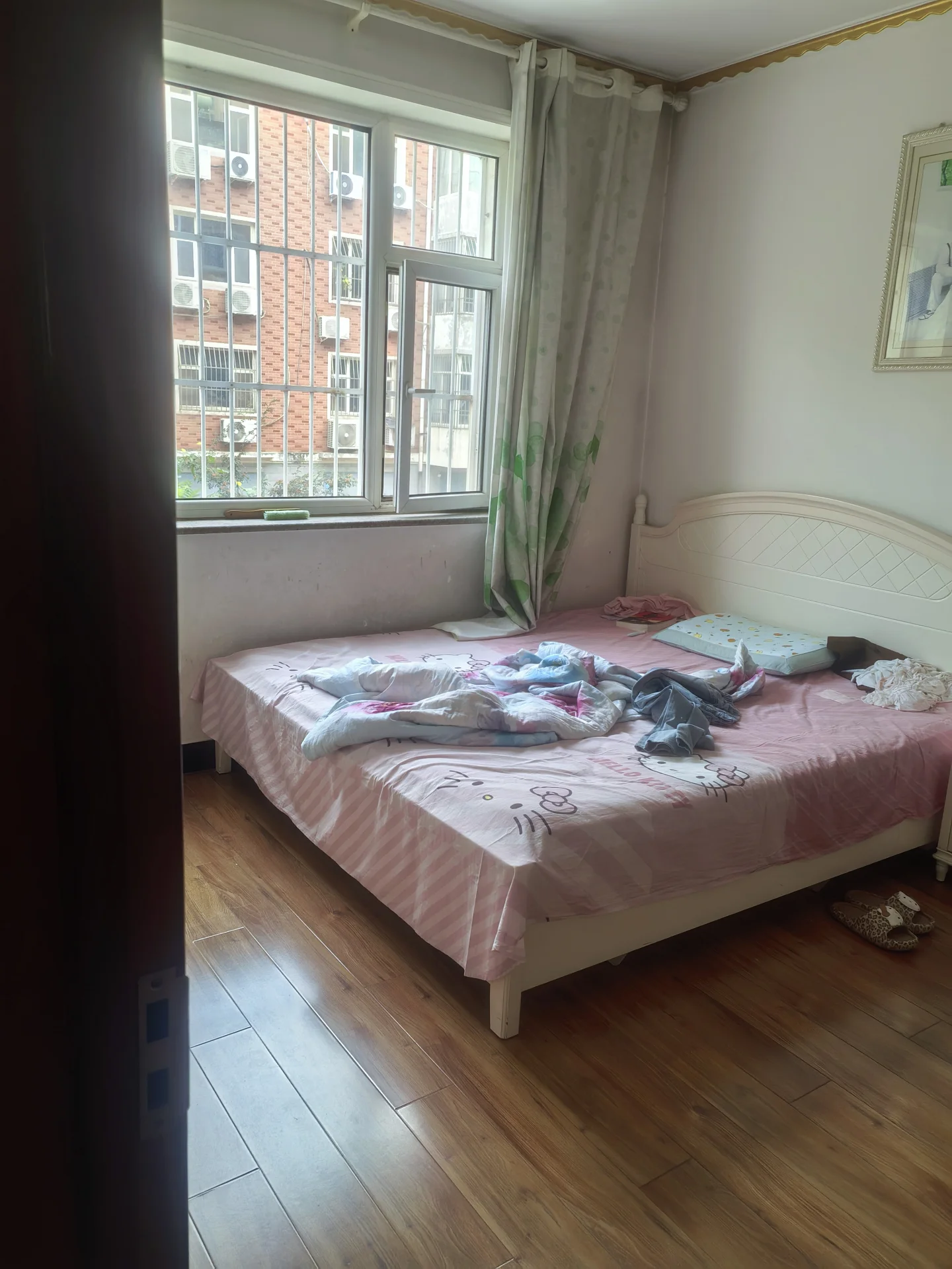 卧室空间，床铺为白色实木框架，床上铺设粉色系床品。房间采光良好，窗外可见城市建筑群，整体装修风格偏向温馨实用。