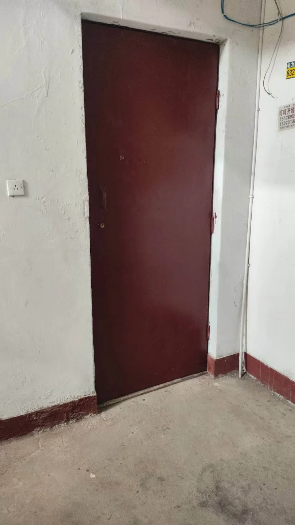 一扇深红色金属门安装在白色墙体上，地面为水泥材质，整体环境简洁，属于建筑的入口或通道。