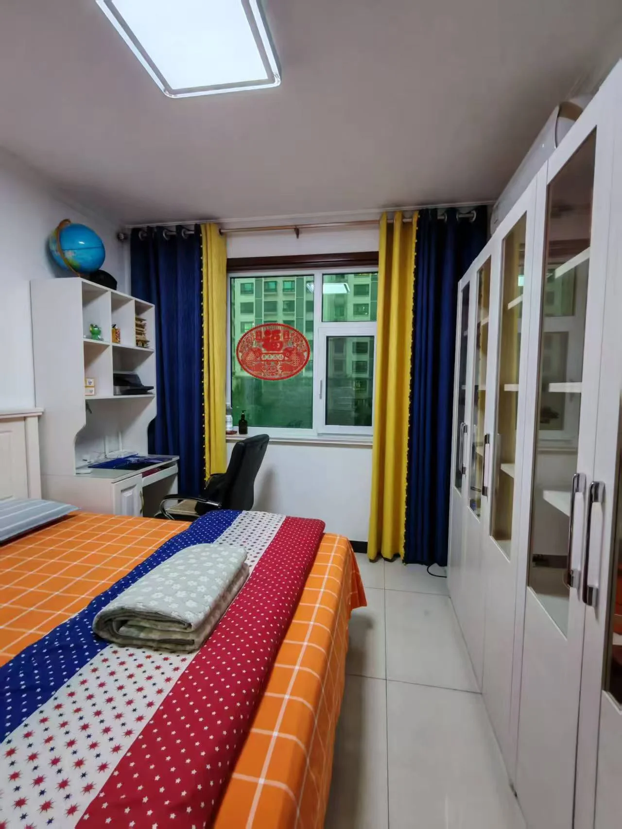 卧室空间，床铺采用多色几何图案床单，配有白色衣柜和简约的家具陈设。房间采光良好，整体装修风格偏向基础实用型。