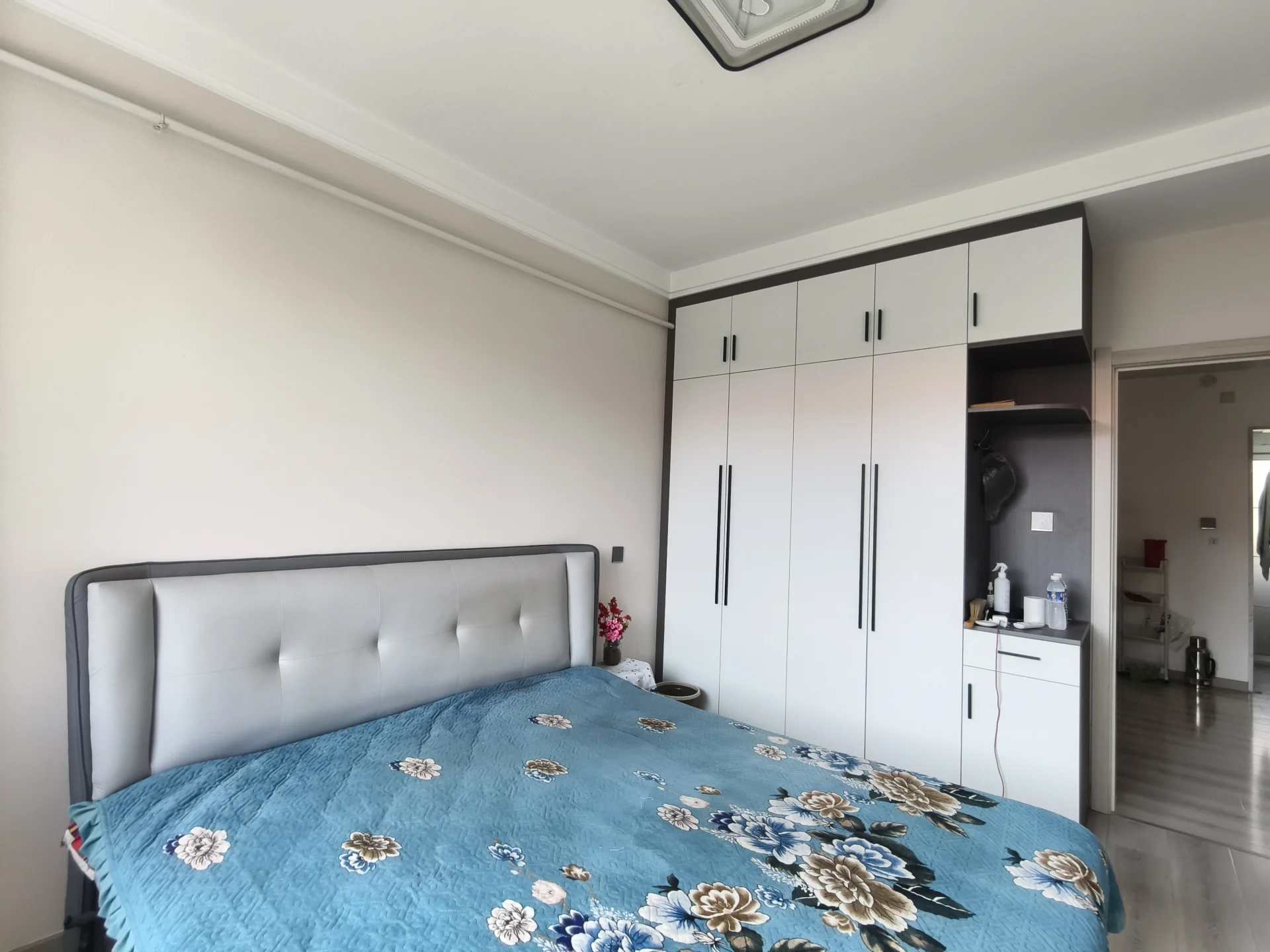 卧室空间，床头采用灰色软包设计，搭配带有蓝色花卉图案的床品。房间整体装修风格偏向现代简约，采光良好，环境整洁舒适。