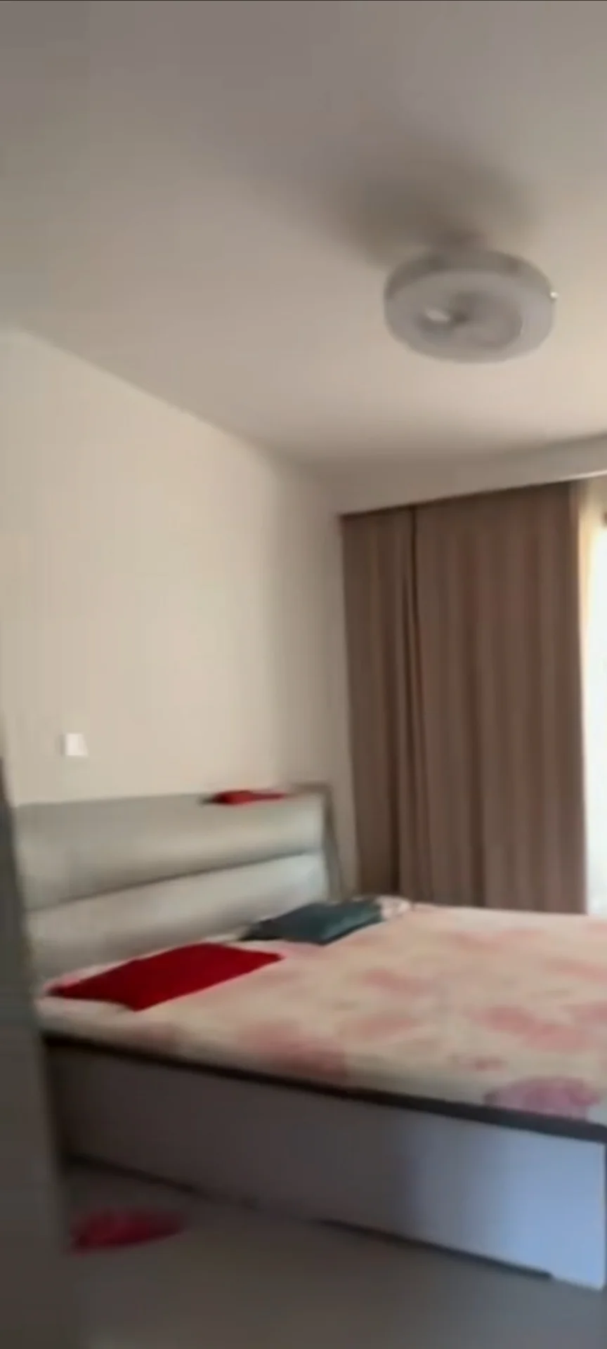 卧室空间，床铺为双人床，搭配粉色系图案床品和米色床架。房间墙面简洁，采光适中，整体呈现基础的现代简约风格。