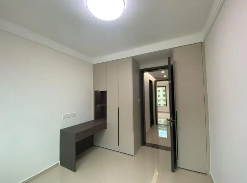 这是一个宽敞的室内空间，墙面为白色，地面为浅色瓷砖。房间内设置了一张深色书桌和一套嵌入式衣柜，整体装修风格简洁实用，采光良好。