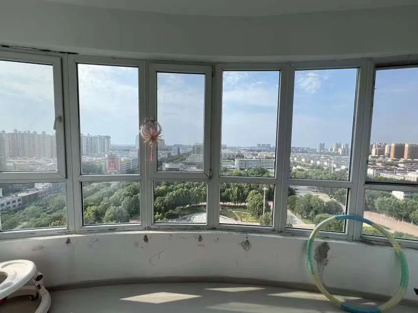 一个带有弧形大窗的室内空间，窗外视野开阔，展现了城市和远处的树木景观。室内装修简洁，光线充足。