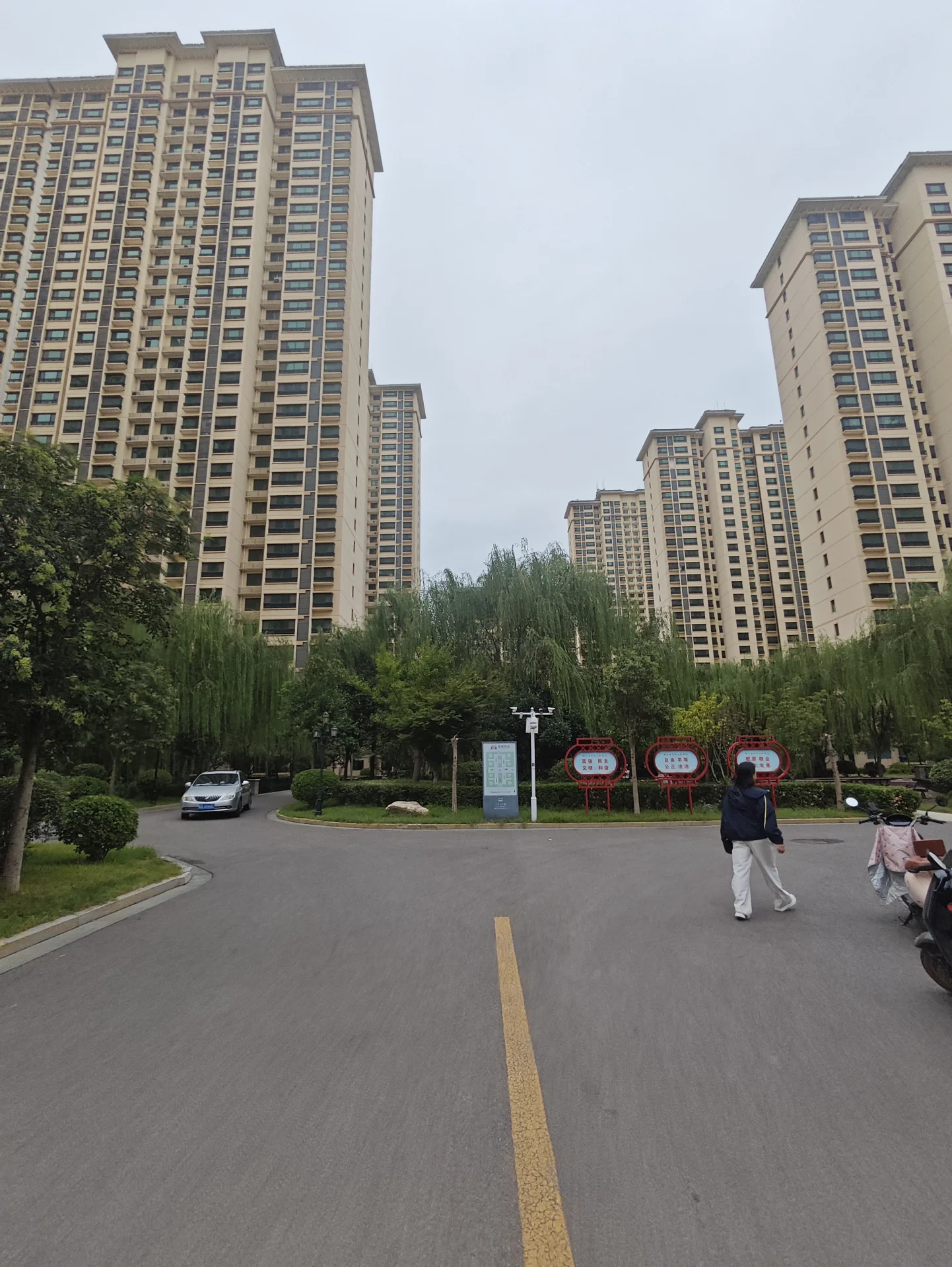 一条宽阔的沥青道路，两侧是高层现代风格住宅楼，道路中央有清晰的黄色标线。背景中绿化带茂密，整体呈现城市居住区的环境。