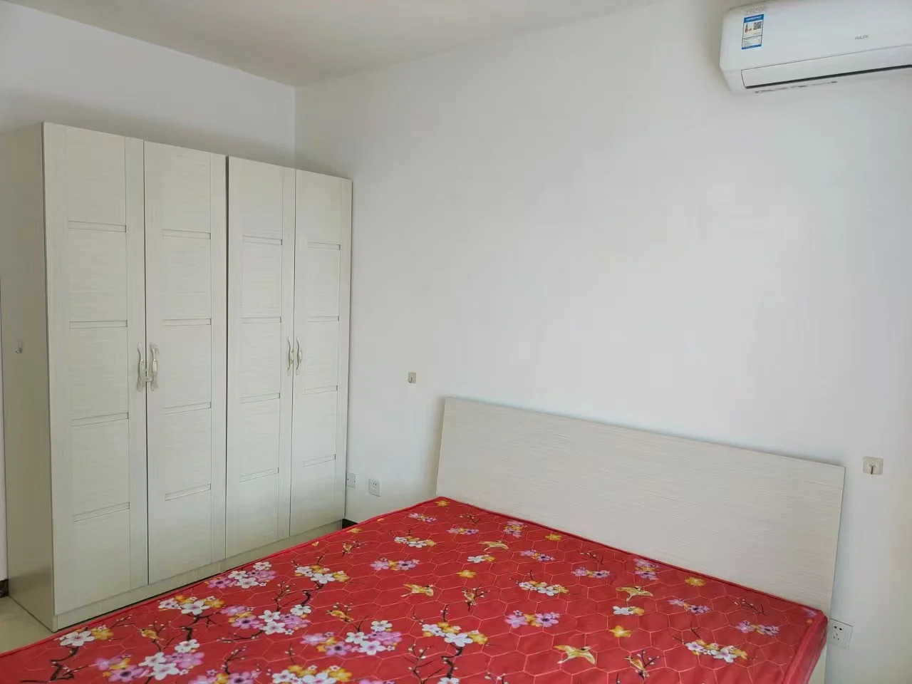 卧室空间，配备白色衣柜和床头板，床铺采用红色花纹床单装饰。房间墙面简洁，采光适中，整体呈现基础的居住环境。