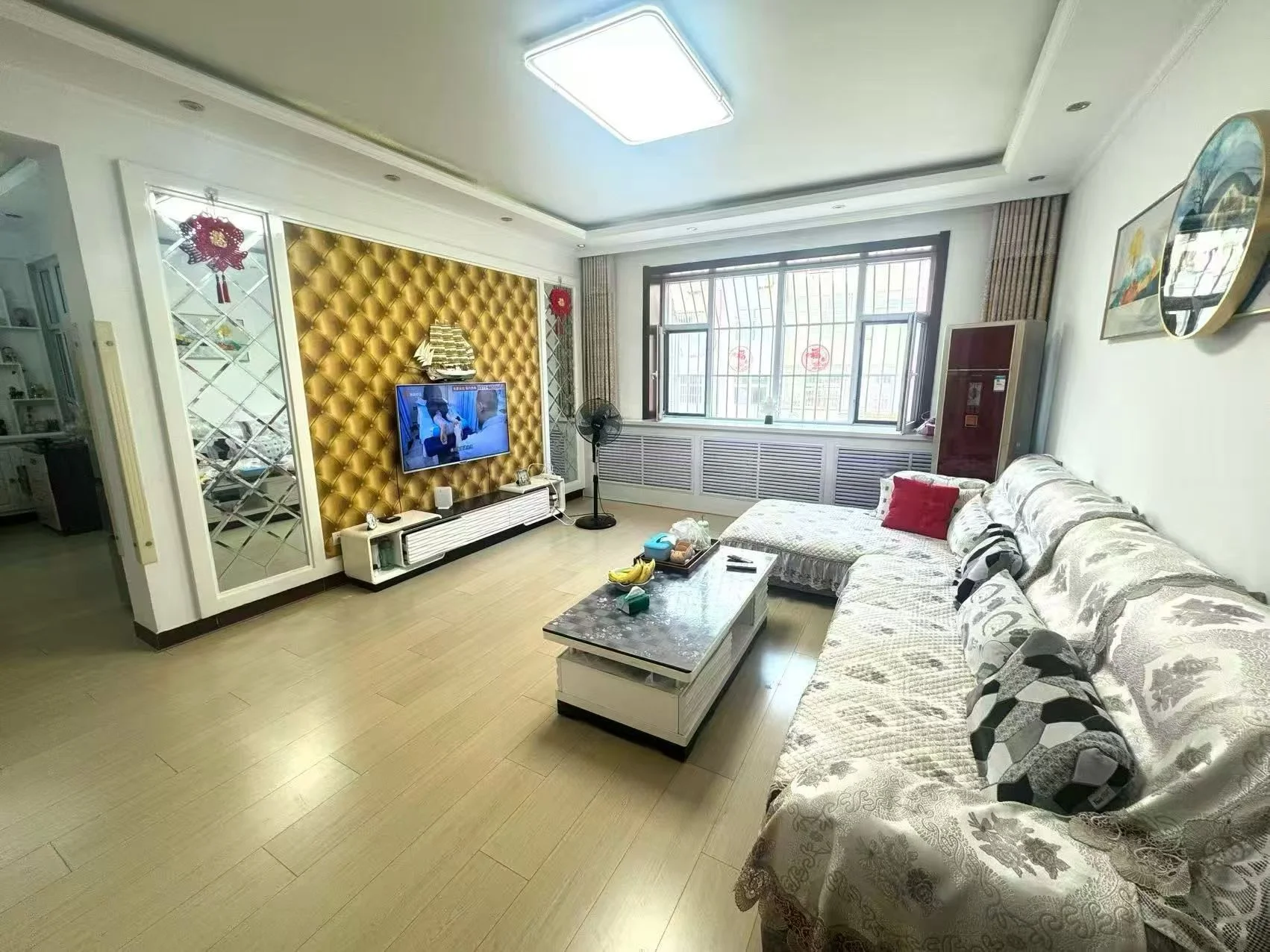 客厅空间，地面铺设浅色木纹地板，墙面部分采用金色菱形纹理装饰，配有电视和简约茶几。采光良好，整体装修风格偏向现代舒适型。