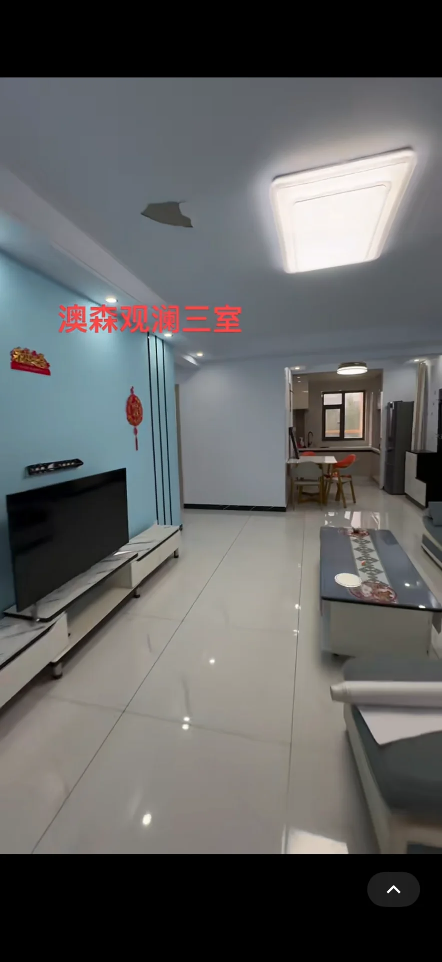 客厅空间，地面铺设大尺寸白色瓷砖，墙面采用浅蓝色调，配有大型电视和简约家具。天花板设计有嵌入式照明，整体采光良好，呈现现代简洁的装修风格。