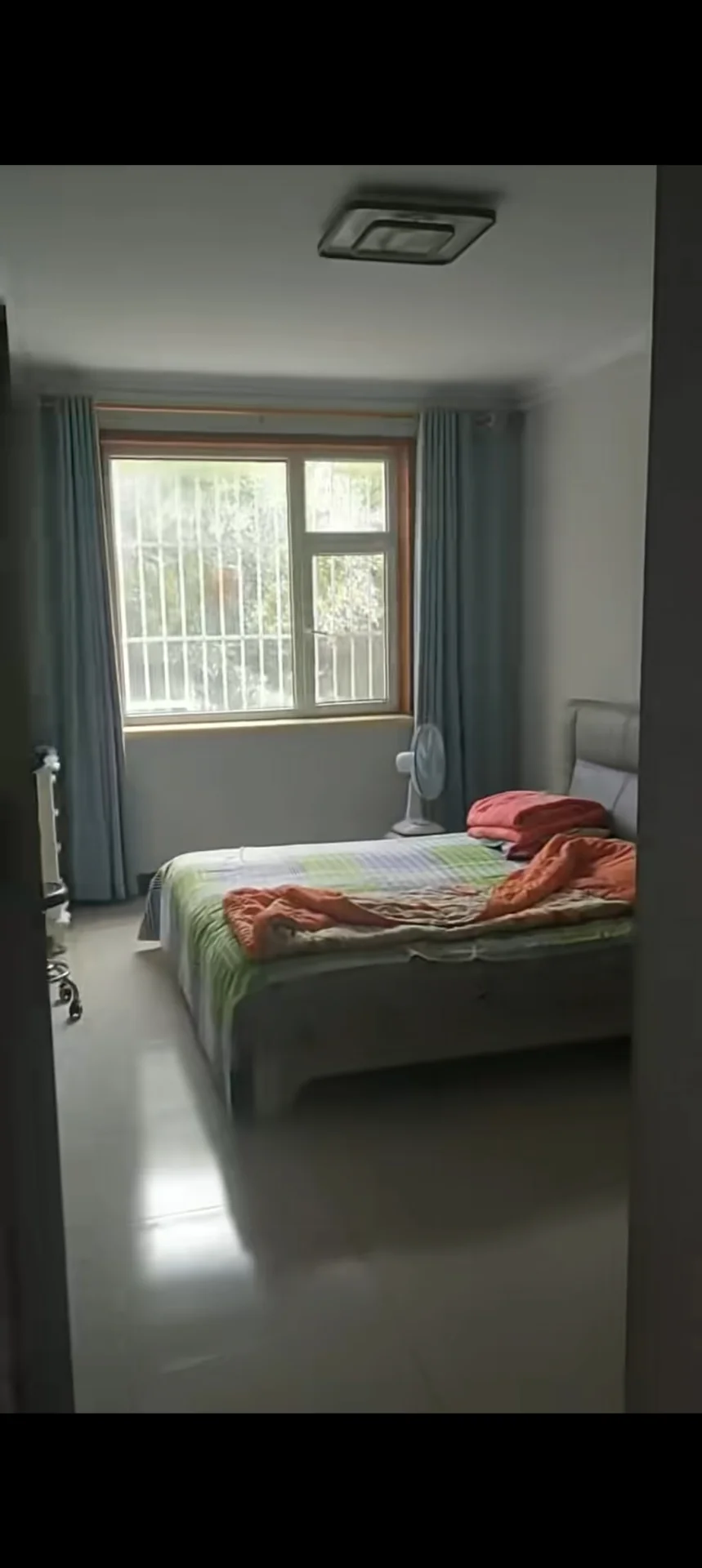 卧室空间，墙面和天花板为白色，配有大型窗户引入自然光。房间内设有一张床和一台风扇，整体装修风格简洁实用，采光良好。
