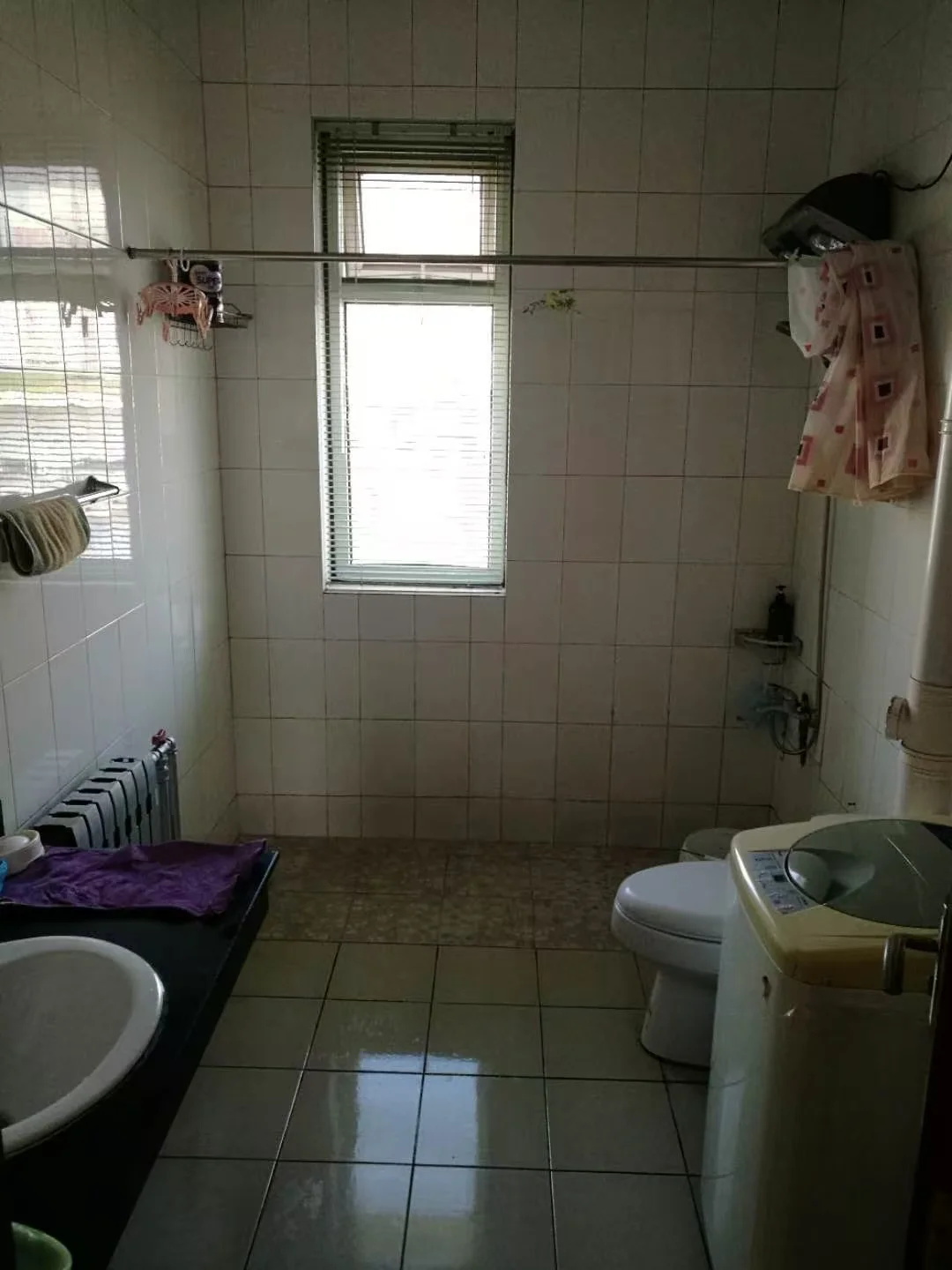卫生间空间，墙面和地面均铺设白色或浅色瓷砖，配有洗手台、马桶和洗衣机等基础卫浴设施。采光适中，整体风格朴素实用。
