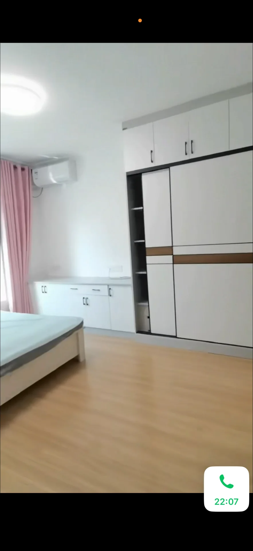 卧室空间，墙面为白色，搭配浅色木纹地板和粉色窗帘。房间内摆放有床和嵌入式衣柜，整体装修风格简洁明快，采光良好。