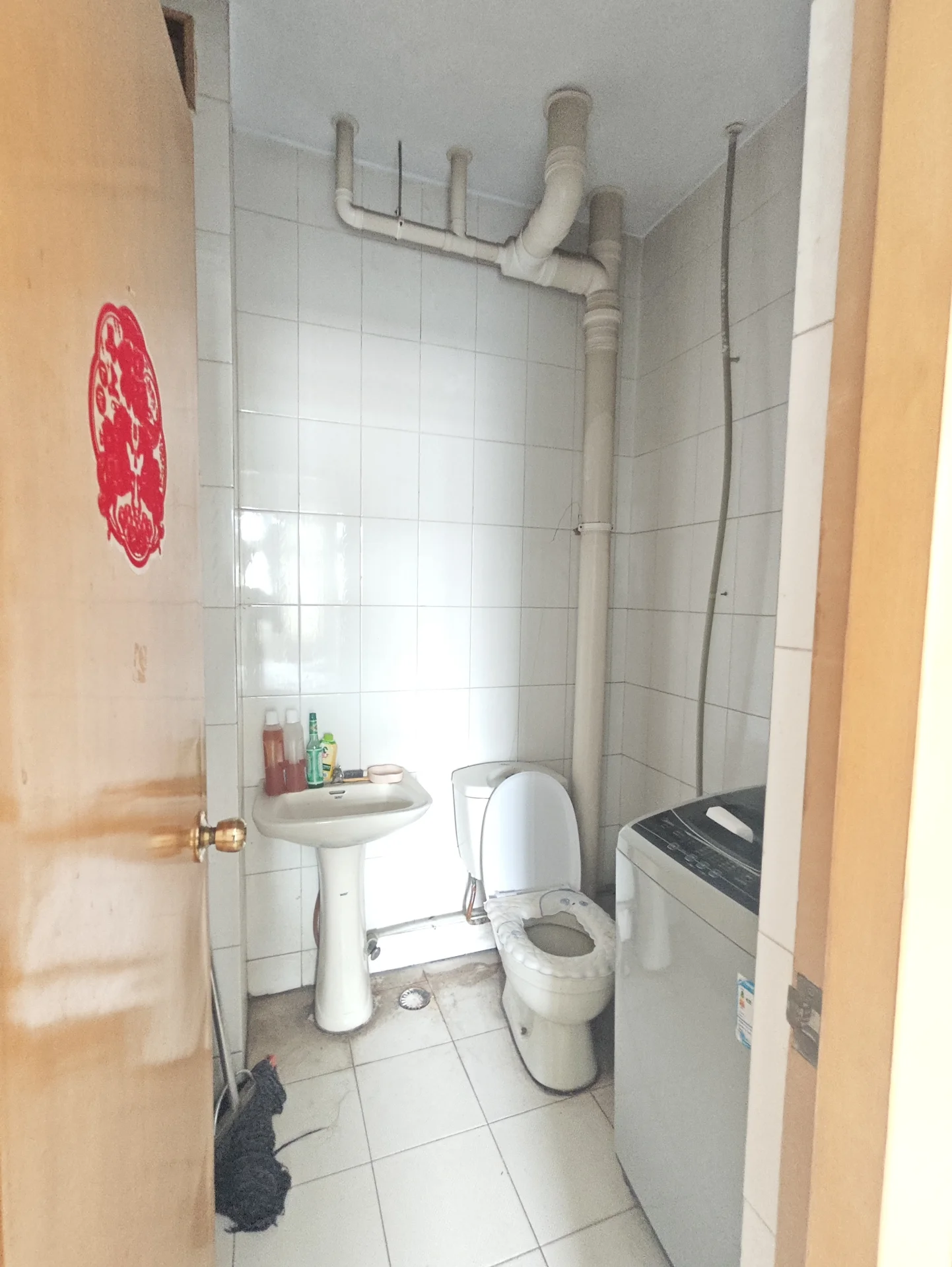 卫生间空间，墙面和地面铺设白色瓷砖，配有白色洗手台、马桶和洗衣机。整体装修风格基础实用，光线明亮。