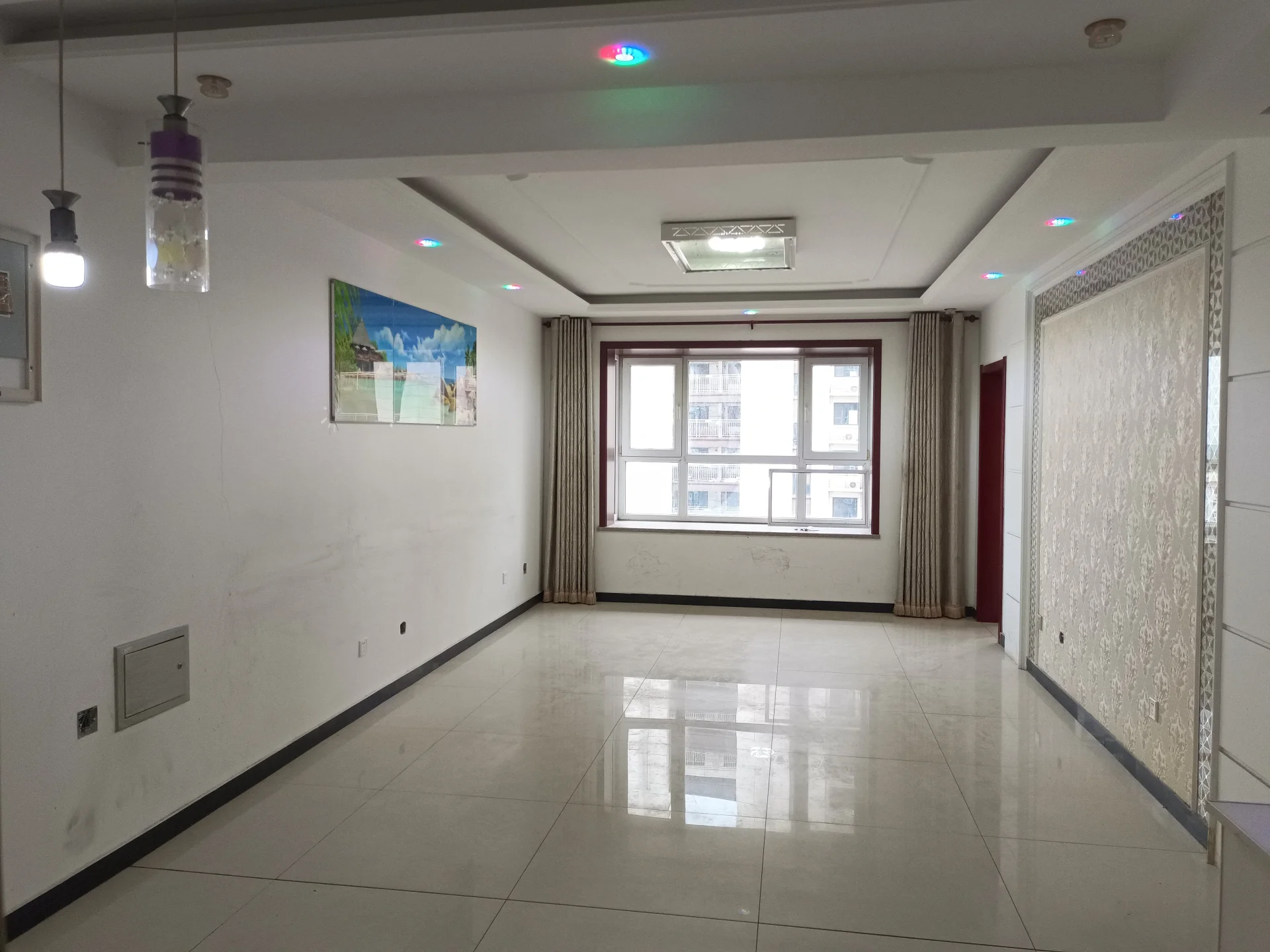 这是一个空旷的室内空间，地面铺设光洁的浅色瓷砖，墙面为白色，配有吊灯照明和一扇窗户。整体环境简洁明亮，呈现出待装修或新交付的商业/居住空间特征。