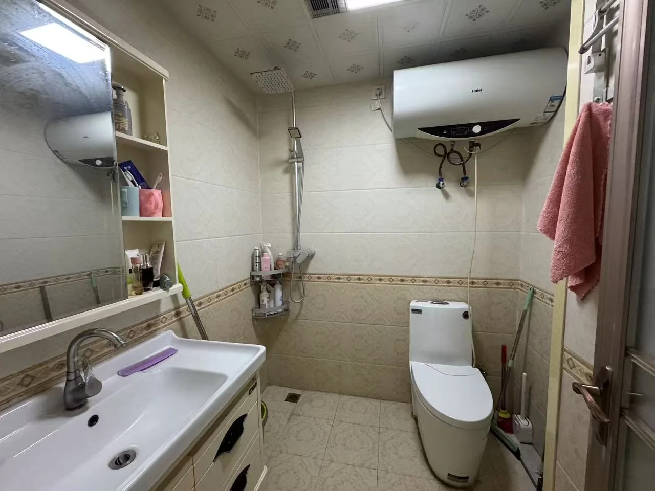 卫生间内部，墙面和地面铺设米色瓷砖，配有白色洗手台、马桶及淋浴区。空间布局功能明确，光线适中，整体风格实用简洁。