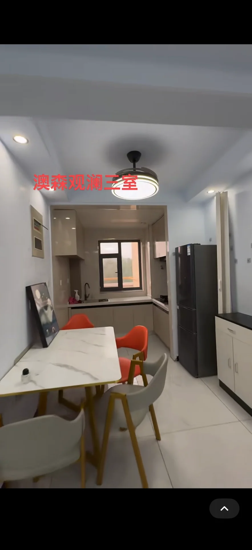 开放式厨房与餐厅结合的室内空间，采用浅色调瓷砖铺设地面，配备白色餐桌和现代风格的餐椅。厨房区域设有嵌入式电器和简洁的橱柜设计，整体采光良好，呈现出现代简约的居住氛围。